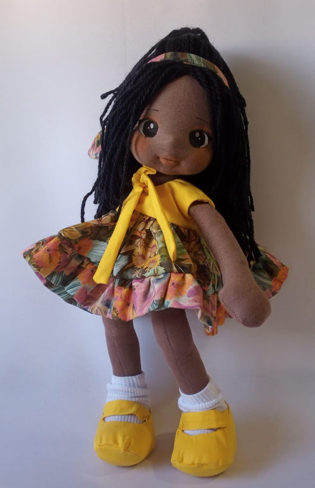 Boneca de pano negra com vestido florido alegre e sapatos amarelos, cabelo preto.