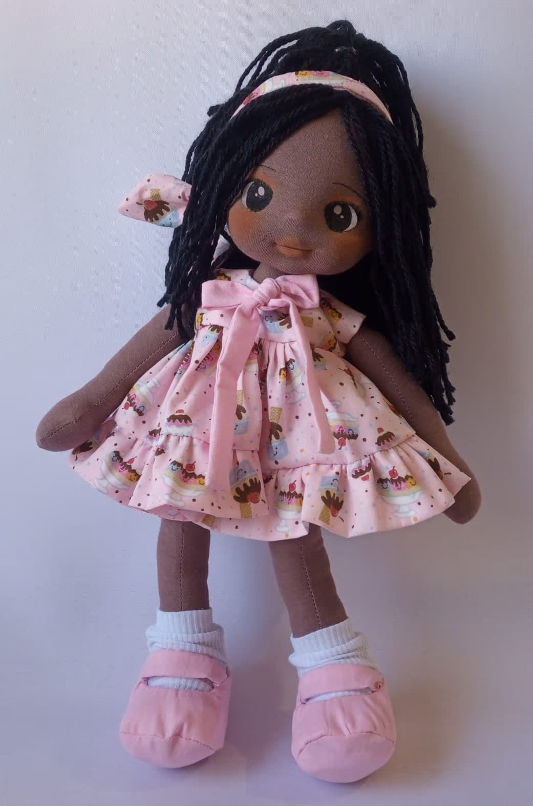 Boneca de pano negra com vestido rosa de docinhos, sapatos rosa e cabelo preto.