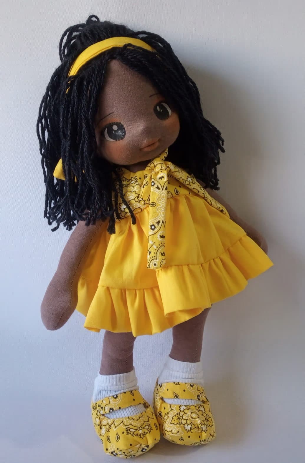 Boneca de pano negra com vestido amarelo e sapatos amarelos, cabelo preto